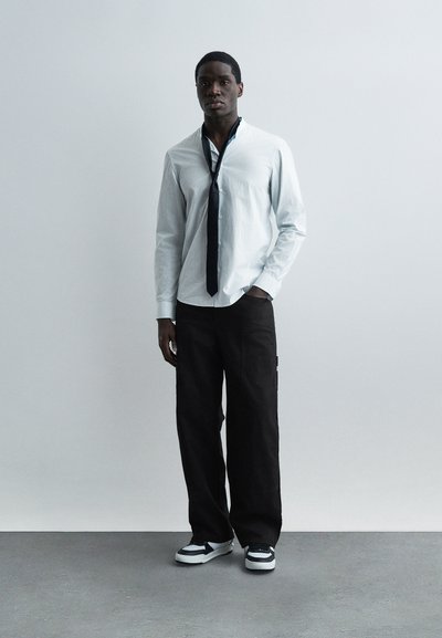 Άνδρας που φοράει ανοιχτό γαλάζιο πουκάμισο με κουμπιά, χαλαρή μαύρη γραβάτα, μαύρο παντελόνι με φαρδιές πατούσες και ασπρόμαυρα αθλητικά παπούτσια, στέκεται μπροστά σε έναν απλό τοίχο.