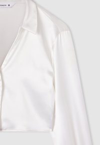 Camicia corta in satin bianco con collo a camicia, maniche lunghe e texture liscia. Presenta un design pulito e una vestibilità su misura.