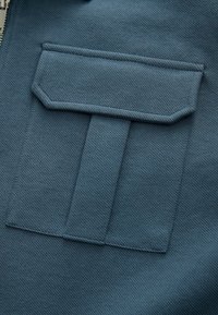 Donker petroleumkleurige stof dichtbij gefotografeerd met een rechthoekige zak met een omgevouwen klep, een textuurrijke weving en nette stikseldetails.