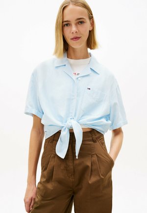 Joven con cabello rubio lacio vistiendo una camisa de rayas azul claro anudada sobre una camiseta blanca y pantalones marrones de tiro alto.