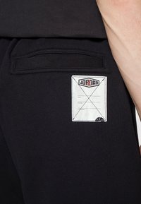 Černá kapsa kalhot s bílou našitou štítkem zobrazující logo Jordana, řádky pro jméno, pozici, hřiště a malý grafický motiv basketbalového míče.