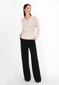 Beżowy sweter dziewiarski z dekoltem w kształcie V i teksturowanymi wzorami, zestawiony z luźnymi czarnymi spodniami i czarnymi butami. Prosty design, luźny krój.