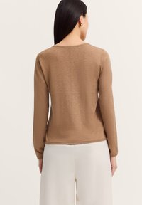 Frau mit langen dunklen Haaren trägt einen beigen Langarmpullover und weiße Hosen, von hinten auf einem schlichten hellen Hintergrund gezeigt.