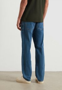 Jeans in denim blu con una vestibilità dritta, caratterizzati da tasche posteriori doppie e una leggera sbiaditura, abbinati a sneakers beige su un pavimento grigio chiaro.