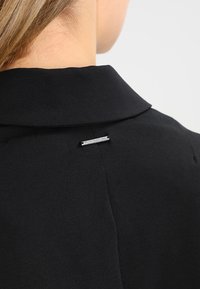 Czarna odzież z kołnierzykiem, z małym metalowym akcentem z tyłu, blisko dekoltu. Tkanina ma gładką, matową fakturę.