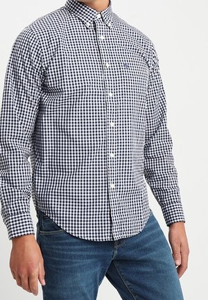 Man wearing a black and white checkered button-up shirt and blue jeans, standing with hands relaxed by sides.