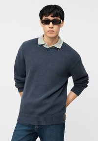 Νεαρός άνδρας που φοράει μαύρα ορθογώνια γυαλιά ηλίου, σκούρο μπλε πλεκτό πουλόβερ πάνω από ανοιχτοπράσινο πουκάμισο με γιακά και μπλε τζιν, στέκεται με τα χέρια πίσω από την πλάτη.