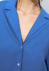 Sininen pellavapaita, jossa on terävä kaulus ja napillinen etuosa. Kangas näyttää teksturoidulta, hieman kiiltävältä ja rentoa istuvuutta edustavalta.