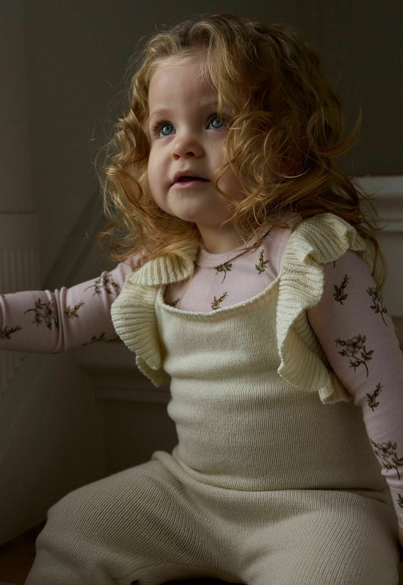 Jeune enfant aux cheveux blonds bouclés portant une chemise fleurie à manches longues et une salopette en tricot crème, regardant vers le haut sous un éclairage doux.