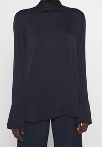 Blusa a maniche lunghe e collo alto in blu navy scuro con finitura testurizzata e polsini svasati. L'orlo è asimmetrico, in modo da completare il taglio rilassato.