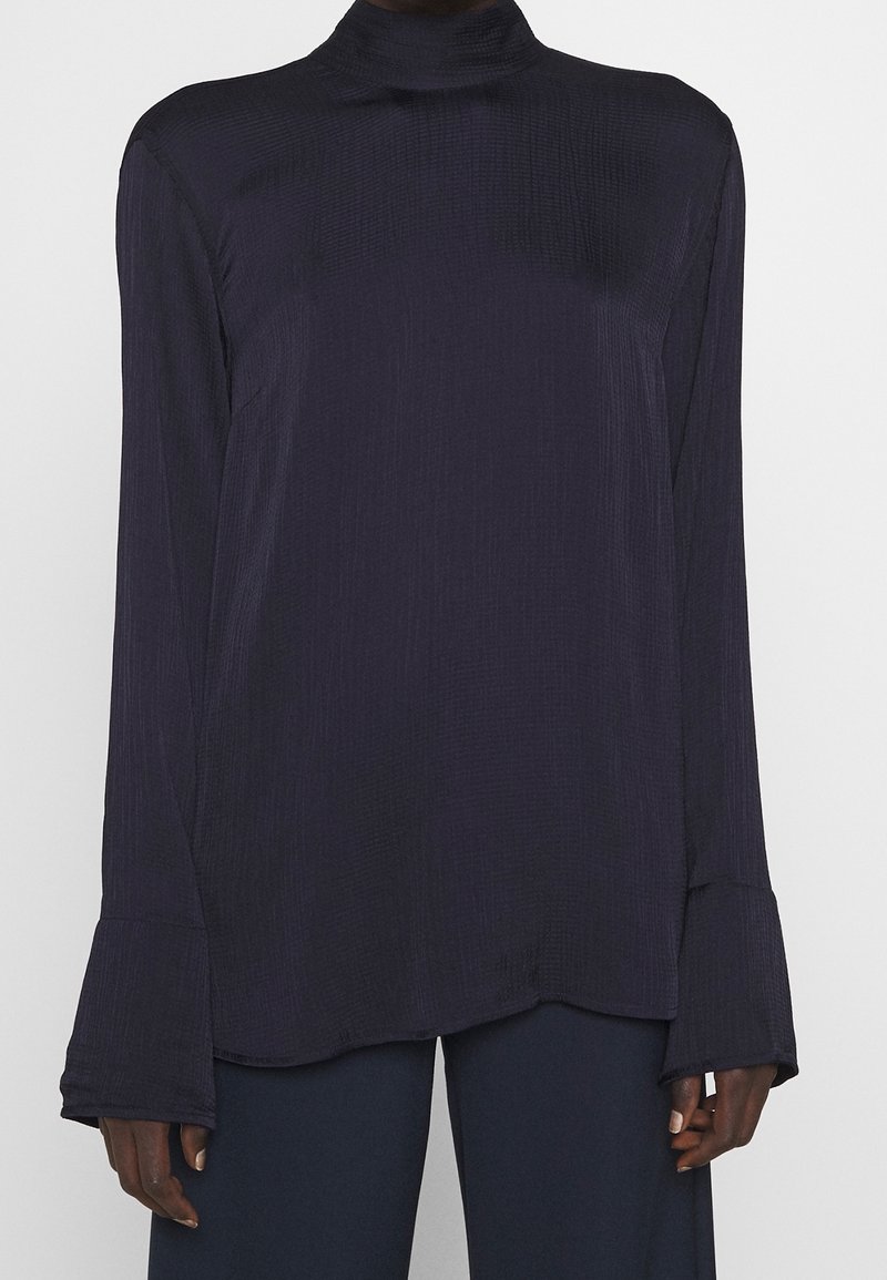 Blusa a maniche lunghe e collo alto in blu navy scuro con finitura testurizzata e polsini svasati. L'orlo è asimmetrico, in modo da completare il taglio rilassato.