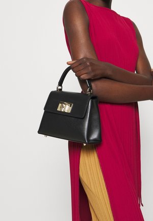 Personne portant une robe rouge sans manches tenant un sac à main noir avec un fermoir doré, debout devant un fond uni clair.