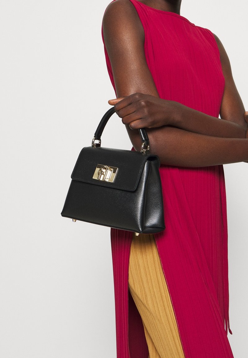 Personne portant une robe rouge sans manches tenant un sac à main noir avec un fermoir doré, debout devant un fond uni clair.
