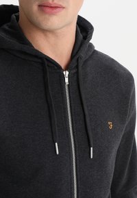 Felpa con zip di colore carbone in tessuto morbido, con zip frontale, cappuccio con cordino e piccolo logo arancione sul petto. Finitura testurizzata.