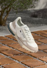 Lacoste sneaker met een bovenwerk van gemengde materialen met witte en beige secties, textuuraccenten en een groene rubberen zool op een betegeld oppervlak.