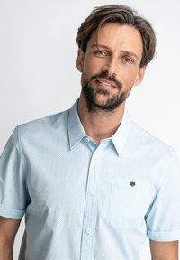 Světle modrá košile s krátkým rukávem, malým límečkem, bílým puntíkovým vzorem a náprsní kapsou s dekoračním knoflíkem.