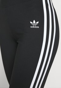 Svarta idrottsleggings med tre vita ränder längs sidan, med en vit Adidas-logotyp som tydligt syns på övre delen av benet.