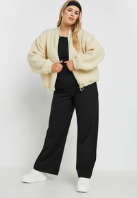 Kremowa, puszysta kurtka bomberka z zamkiem z przodu, szerokimi rękawami i ściągaczem na dole, w połączeniu z czarnymi spodniami w szerokim kroju i białymi sneakersami.