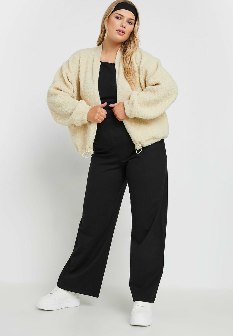 Kremowa, puszysta kurtka bomberka z zamkiem z przodu, szerokimi rękawami i ściągaczem na dole, w połączeniu z czarnymi spodniami w szerokim kroju i białymi sneakersami.
