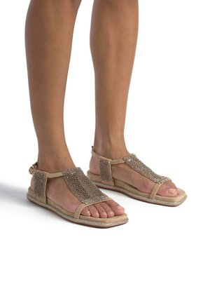 Sandali beige realizzati in materiale morbido, con un design di cinturino testurizzato e decorato con perline e una fibbia per la caviglia regolabile, dotati di una suola piatta.