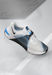 White and black athletic shoe with blue accents, featuring a textured mesh upper, round toe, and a flat rubber sole. Laces and logo visible.