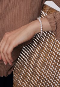 Perłowa bransoletka z błyszczącymi białymi koralikami na smukłym nadgarstku, w brązowym swetrze z prążkami, na tle plecionki z rattanu.