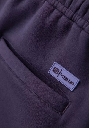 Dunkellila Jogginghosen verfügen über eine genähte Tasche und ein rechteckiges Etikett mit "LEVEL UP" in weißen Buchstaben auf einem strukturierten Stoff.