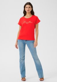 Tricou roșu din bumbac, cu gât rotund și mâneci scurte, având un imprimeu aurie cu litere cursive. Asociat cu blugi deschiși la culoare, evazați.