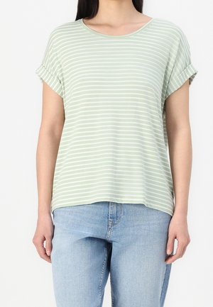 Femme portant une chemise à manches courtes rayée vert clair et blanc avec des poignets retroussés et un jean bleu clair.