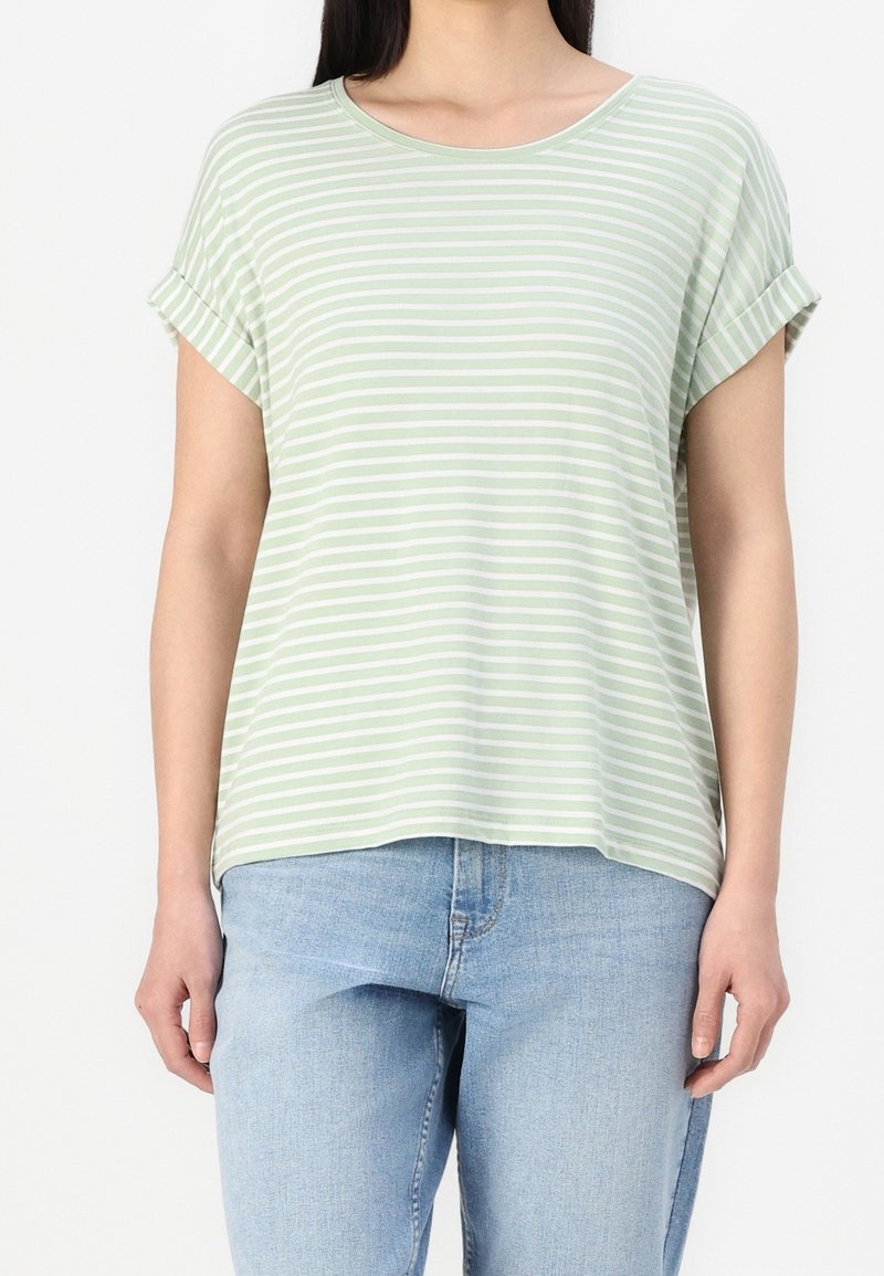 Femme portant une chemise à manches courtes rayée vert clair et blanc avec des poignets retroussés et un jean bleu clair.