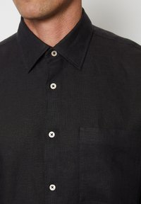 Zwarte linnen shirt met een puntige kraag, voorzien van een enkele borstzak en lichtgekleurde knopen. De textuur lijkt iets ruw.