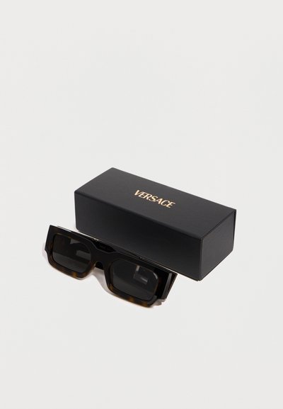 Μαύρα ορθογώνια γυαλιά ηλίου με σκελετό σε χρώμα χελώνας δίπλα σε μαύρη θήκη Versace με χρυσά γράμματα σε λευκό φόντο.