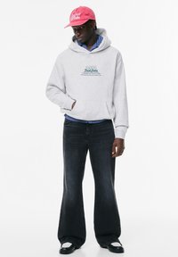 Felpa grigia con testo grafico, strisce blu sotto; abbinata a jeans neri a zampa d'elefante e scarpe nere. Il cappellino rosa presenta dettagli con logo.