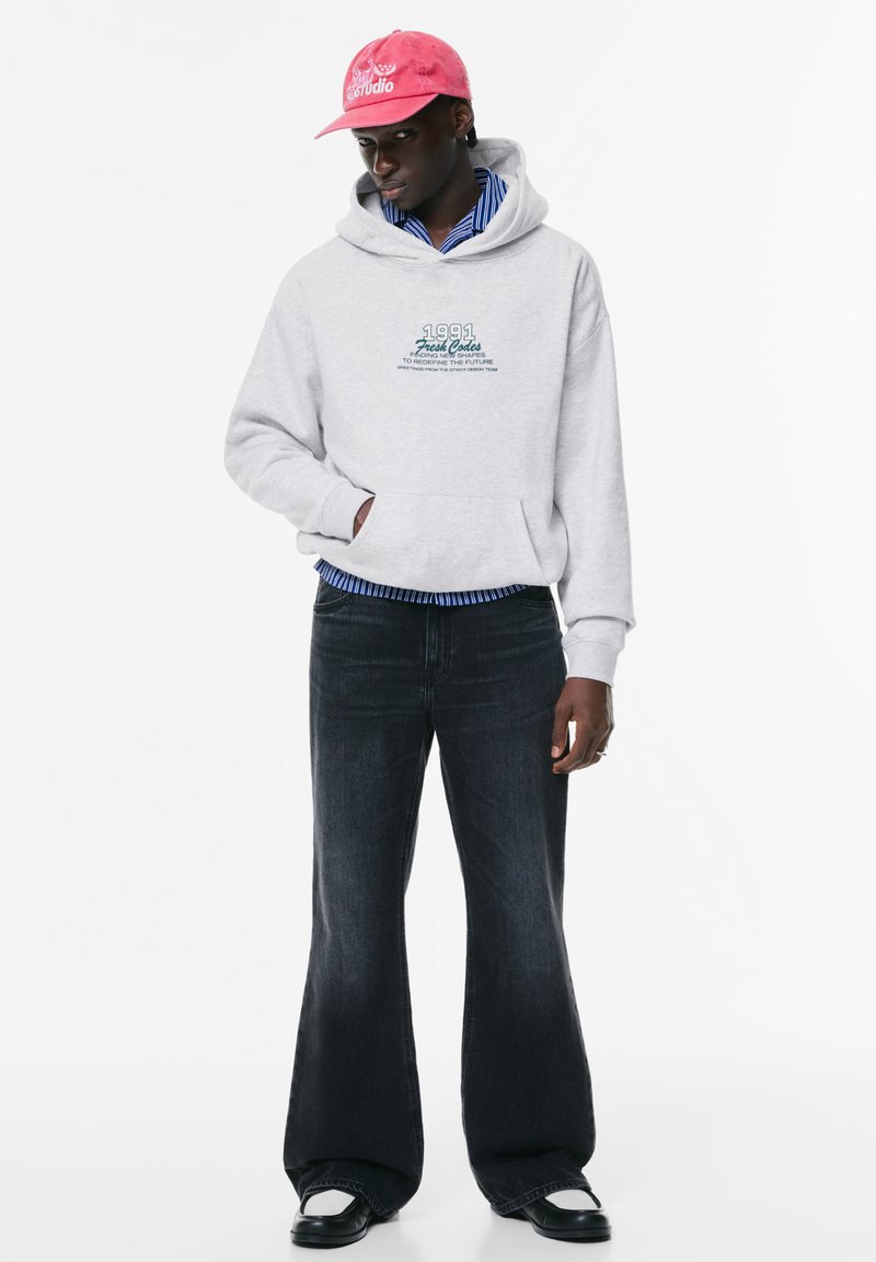 Felpa grigia con testo grafico, strisce blu sotto; abbinata a jeans neri a zampa d'elefante e scarpe nere. Il cappellino rosa presenta dettagli con logo.