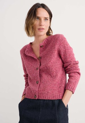 Woman with short brown hair wearing a pink buttoned knit cardigan and dark pants, standing with hands in pockets against a plain background.