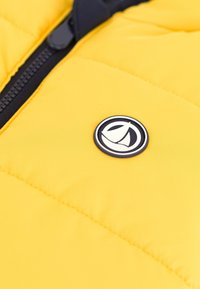 Žuta pufasta jakna sa crnim zipom i okruglim logotipom s dizajnom čamca u bijeloj i crnoj boji. Teksturna površina tkanine.
