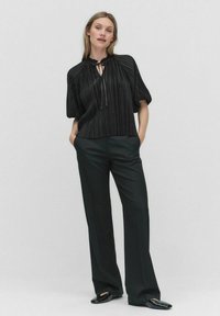 Sort plisseret bluse med båndlukning, korte ærmer, parret med højtaljede sorte bukser og flade, runde sorte sko.