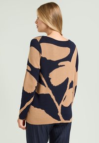 Maglione lavorato a maglia beige e blu scuro con un motivo floreale oversize, polsini a coste e scollo rotondo, abbinato a pantaloni a righe.