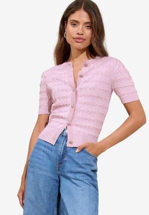 Lyserød, kortærmet cardigan med et struktureret bølgemønster, der har en rund halsudskæring og dekorative knapper, parret med blå denimbukser.