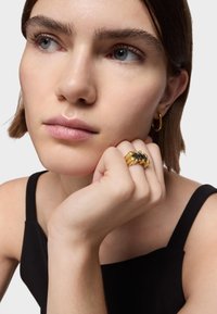 Anillos en tono dorado con acentos negros, acabado suave y formas redondeadas, llevados en una mano cerca de la cara, con un top negro de tirantes finos.