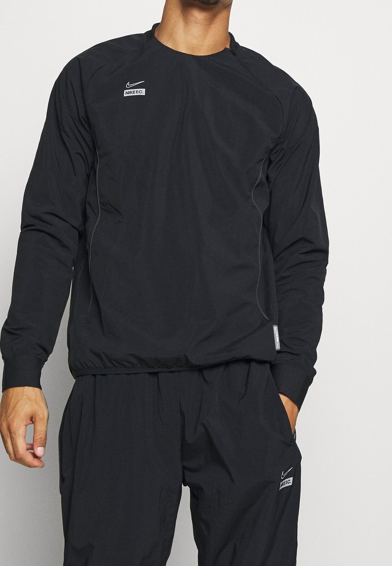 Homme portant une tenue de sport Nike F.C. noire avec un haut à manches longues et un pantalon assorti, debout devant un fond clair uni.