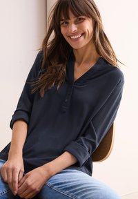 Blusa blu navy a maniche lunghe con scollo a V e tre bottoni. Tessuto morbido, maniche arrotolabili e vestibilità rilassata. Indossata con jeans azzurri.