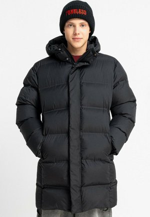 Czarna kurtka puchowa z kapturem, z pikowanym wzorem i zapięciami na napy. Model nosi czarną czapkę z napisem "BEZSENSOWNY" w kolorze czerwonym.