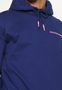 Sudadera marina hecha de una mezcla de algodón, con cordón ajustable con puntas rosas y un logotipo rosa en el pecho. Textura suave y ajuste regular.