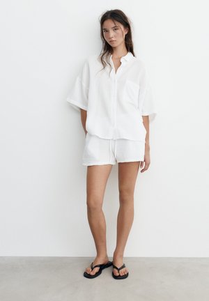 Jeune femme en chemise blanche ample à manches courtes et short assorti, debout pieds nus sur un sol gris contre un mur blanc uni.