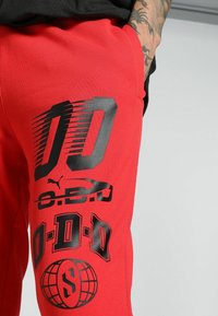 Červené tepláky s veľkými čiernymi grafikami, vrátane čísiel a log, vyrobené z hladkej, elastickej textílie.