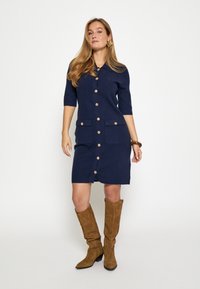 Marineblå skjortekjole med korte ærmer og to forlommer. Har træknapper og en lige pasform. Bruges med tanfarvede knæhøje støvler.