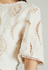 Witte katoenen top met ingewikkelde beige borduursels, kantdetails en golvende randen op de mouwen, versierd met bloem- en geometrische patronen.