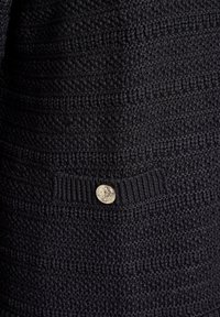 Svart stickad tröja med texturerade mönster, ribbade detaljer och en dekorativ silverfärgad knapp vid fållen.