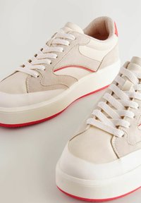 Sneaker beige con finitura liscia e scamosciata, lacci bianchi, dettagli rossi e una suola spessa bianca con fondo rosso.